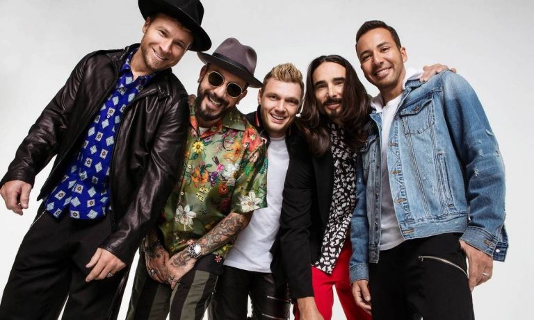 Banda Backstreet Boys posa para foto em fundo branco e com roupas coloridas, rindo e se abraçando