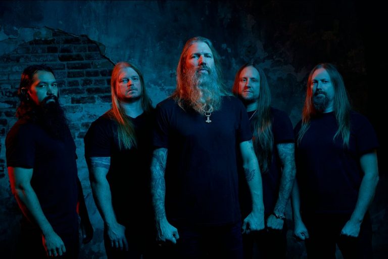 Banda Amon Amarth posa para foto em frente a muro destruido e com luzes vermelhas e azuis